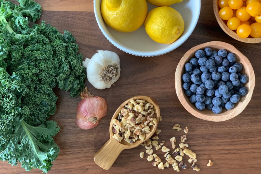 a table decorated with fresh kale, blueberries, tomatoes, walnuts, garlic, lemon and shallot