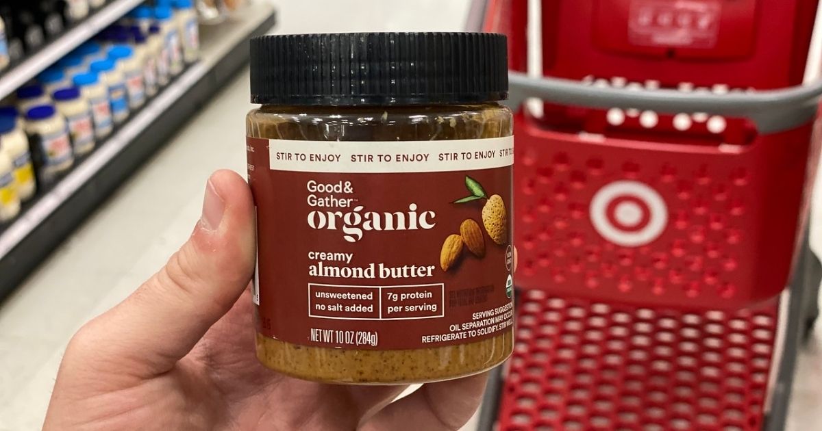 A hand holding a jar of almond butter in front of a Target shopping cart