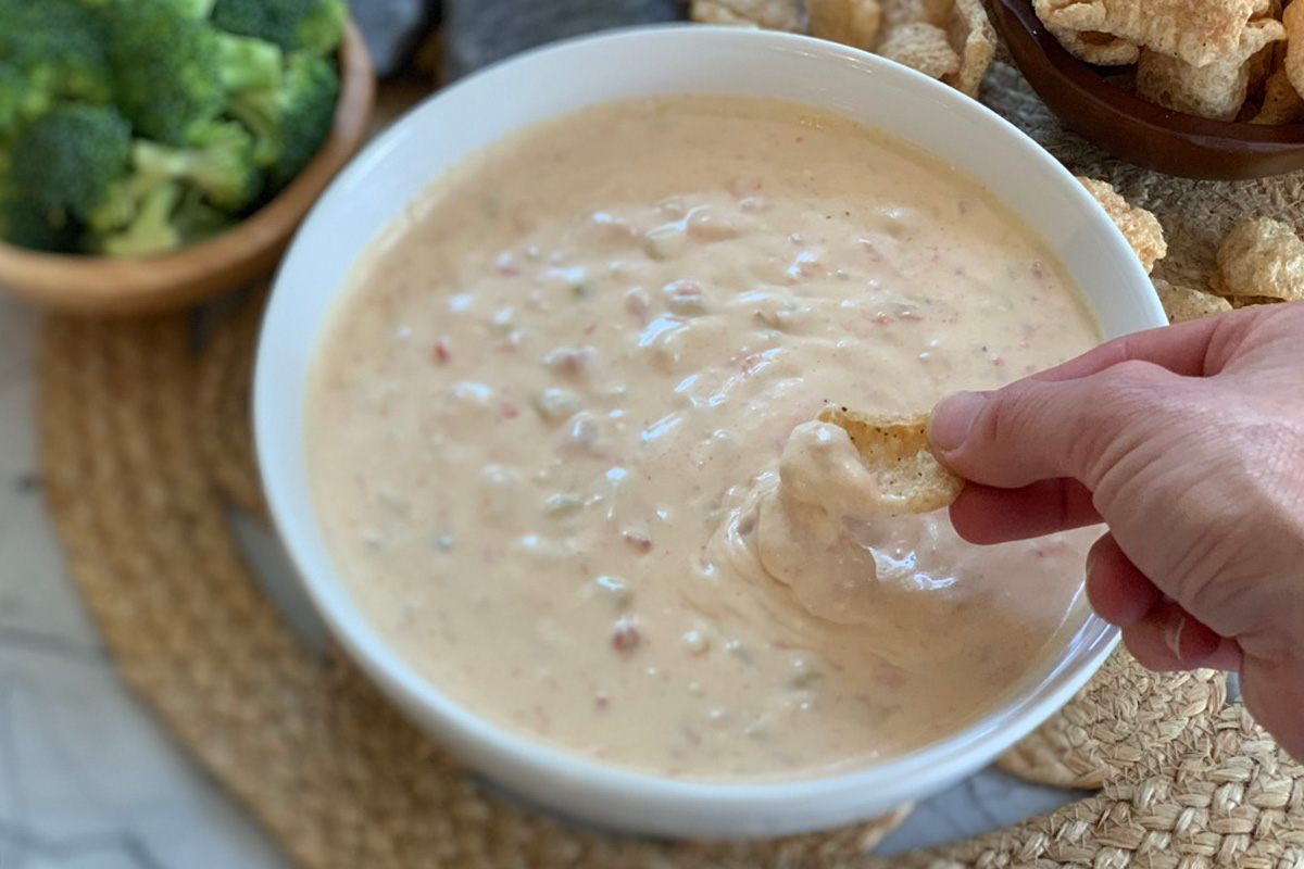 a hand dipping a pork rind into creamy queso dip