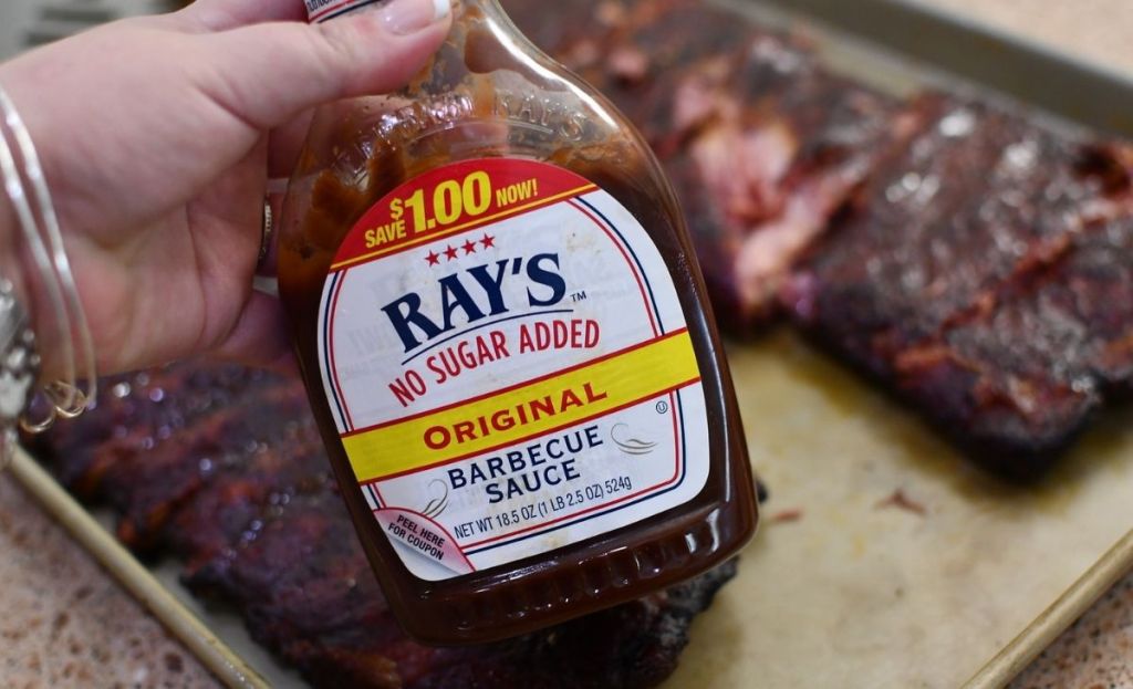 A hand holding a bottle of BBQ sauce in front of ribs