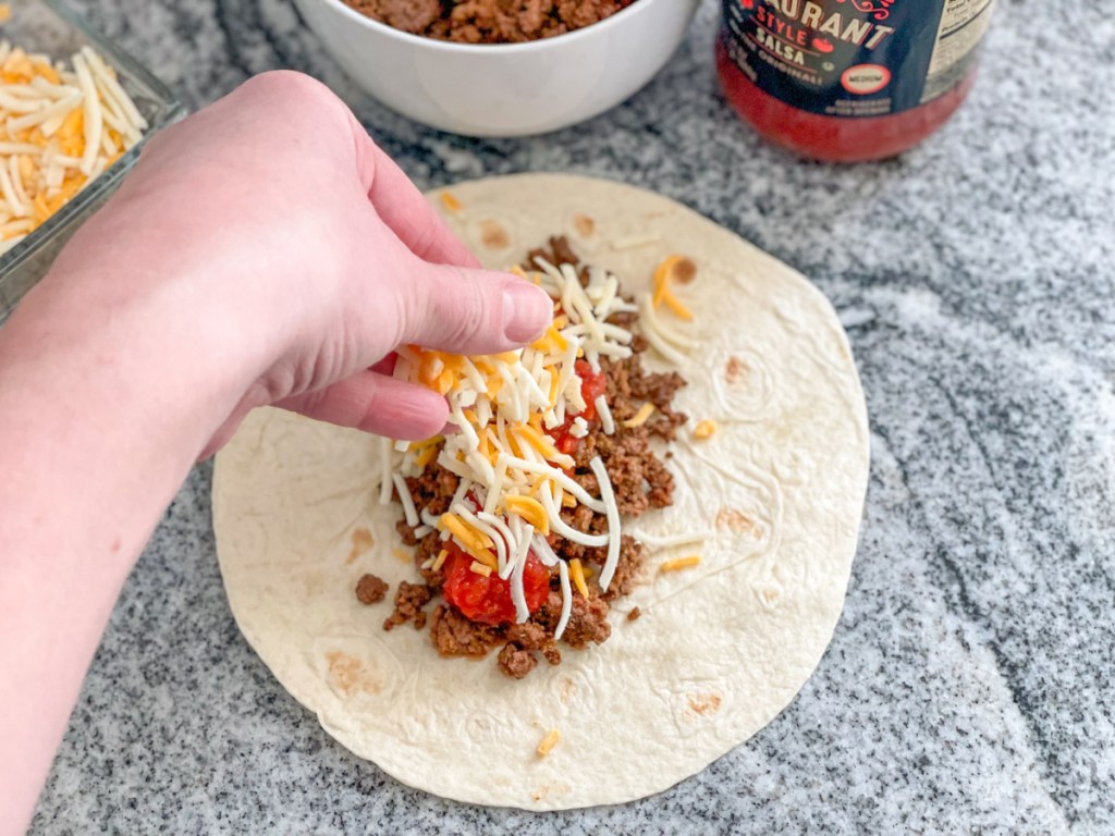 adding shredded cheese to a tortilla with ground beef and salsa