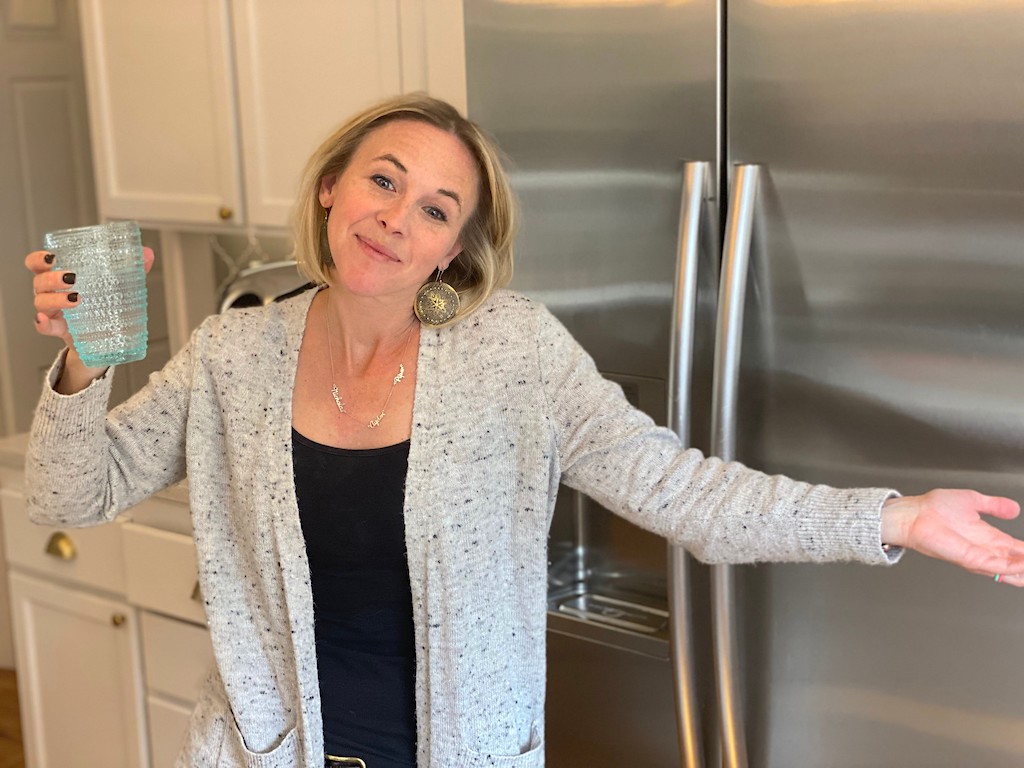 woman holding glass of water by fridge with arms out 