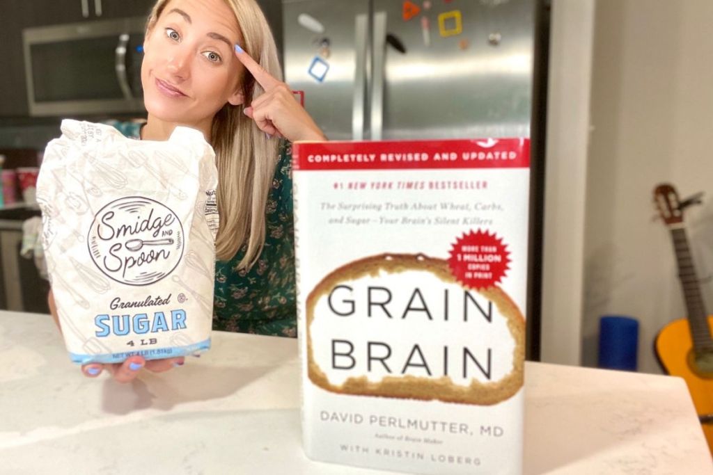 A woman holding a bag of sugar next to a hardcover of Grain Brain
