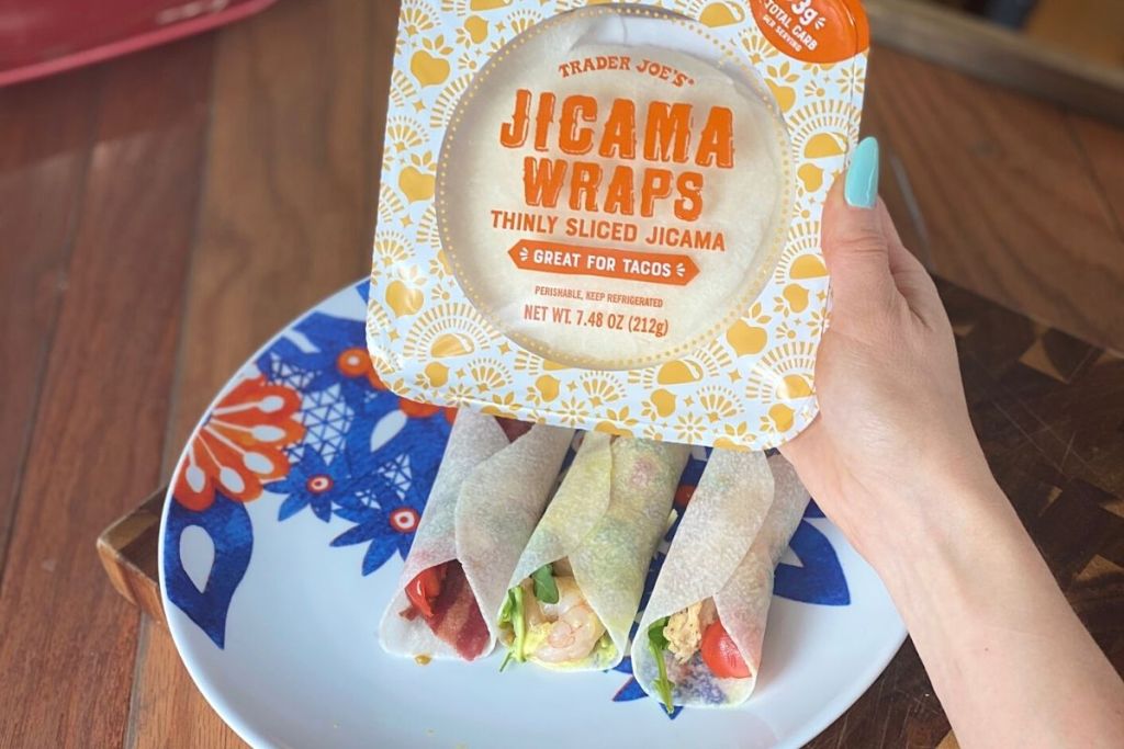 A hand holding a package of jicama wraps next to a plate of tacos