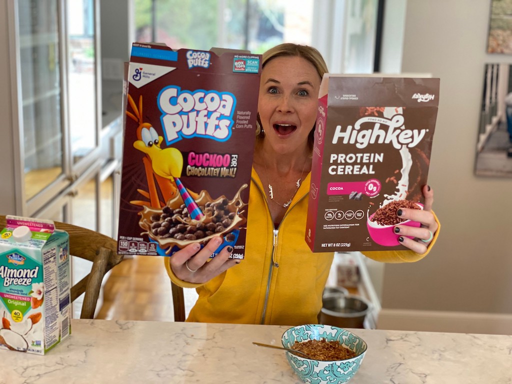 woman holding up Cocoa Puffs and HighKey protein cereal boxes 