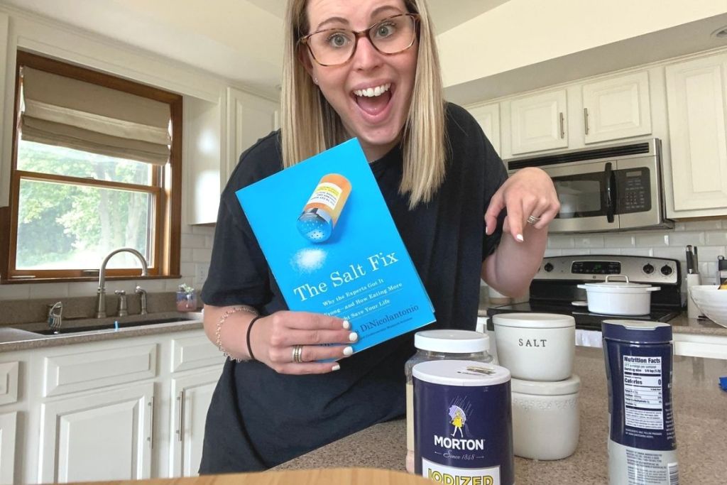 A woman holding a book and pointing to containers of salt on the counter