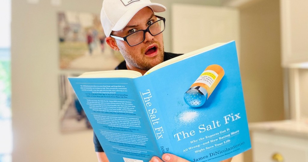 man wearing black glasses and white hat holding the salt fix book