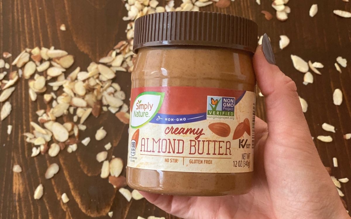 A hand holding a jar of almond butter over a table with some almonds on it