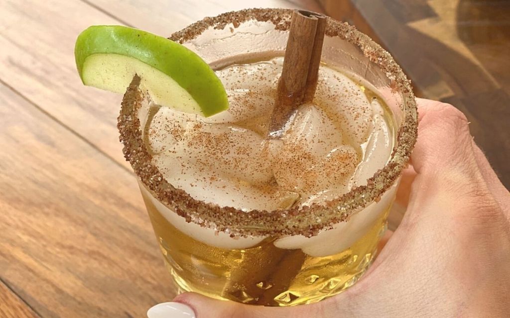 A hand holding a drink with cinnamon sticks and an apple slice on the rim