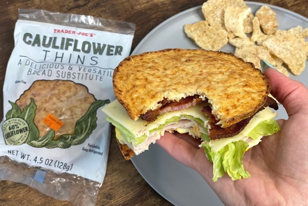 A hand holding a keto sandwich next to a package of cauliflower thins on a table
