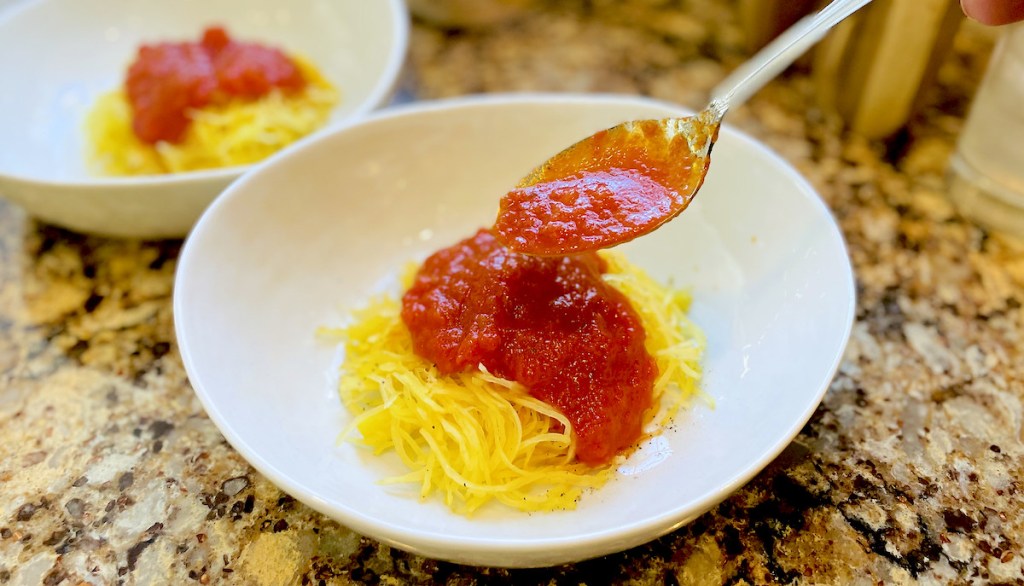stainless steel spoon putting red marinara sauce on top of spaghetti squash in white bowl
