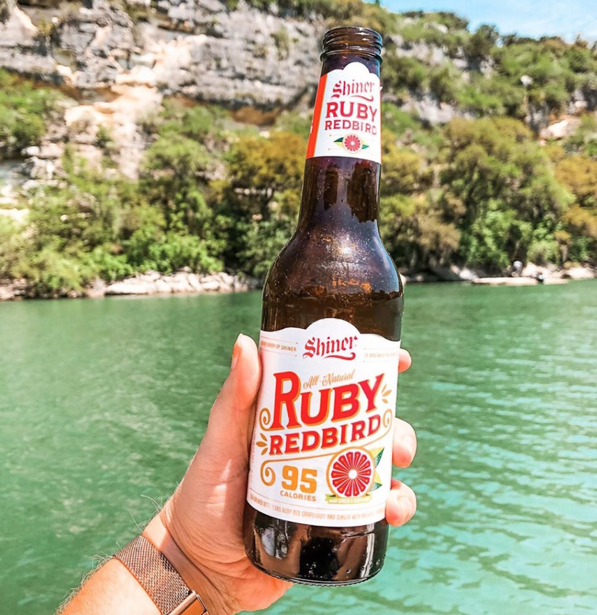 hand holding a glass bottle of shiner ruby redbird low carb alcoholic beverage in front of blue lake
