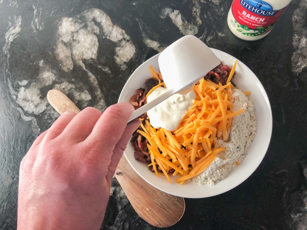 pouring ranch dressing into bowl with bacon, shredded cheddar, and ranch packet