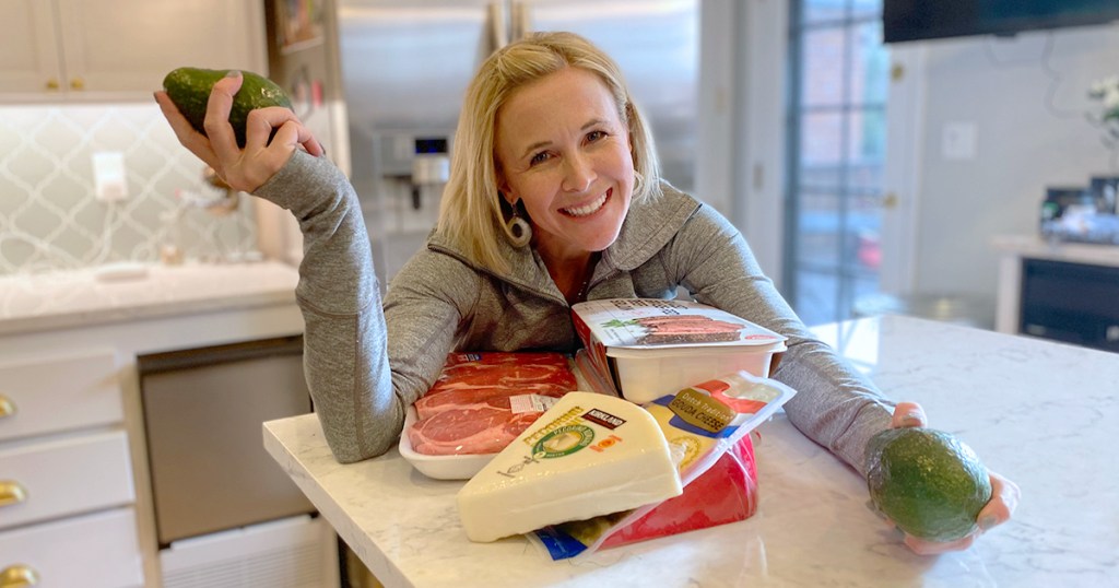 woman leaning on kitchen counter holding avocados with steaks and blocks of cheese