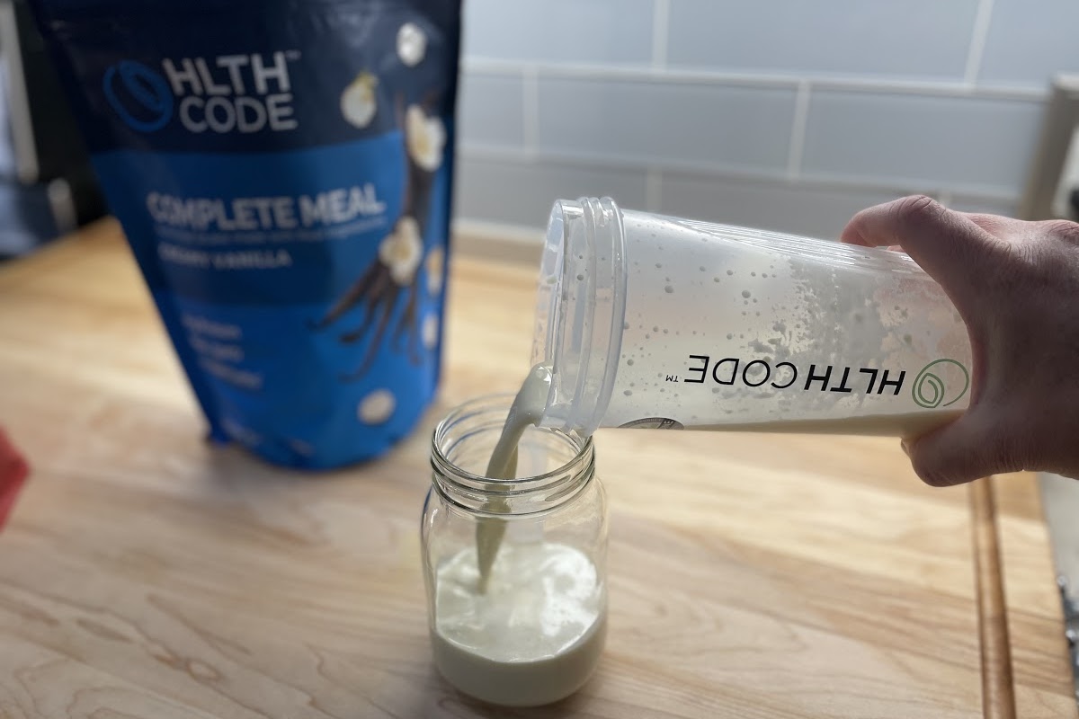 hand pouring vanilla meal replacement drink into tumbler on counter with bag in background