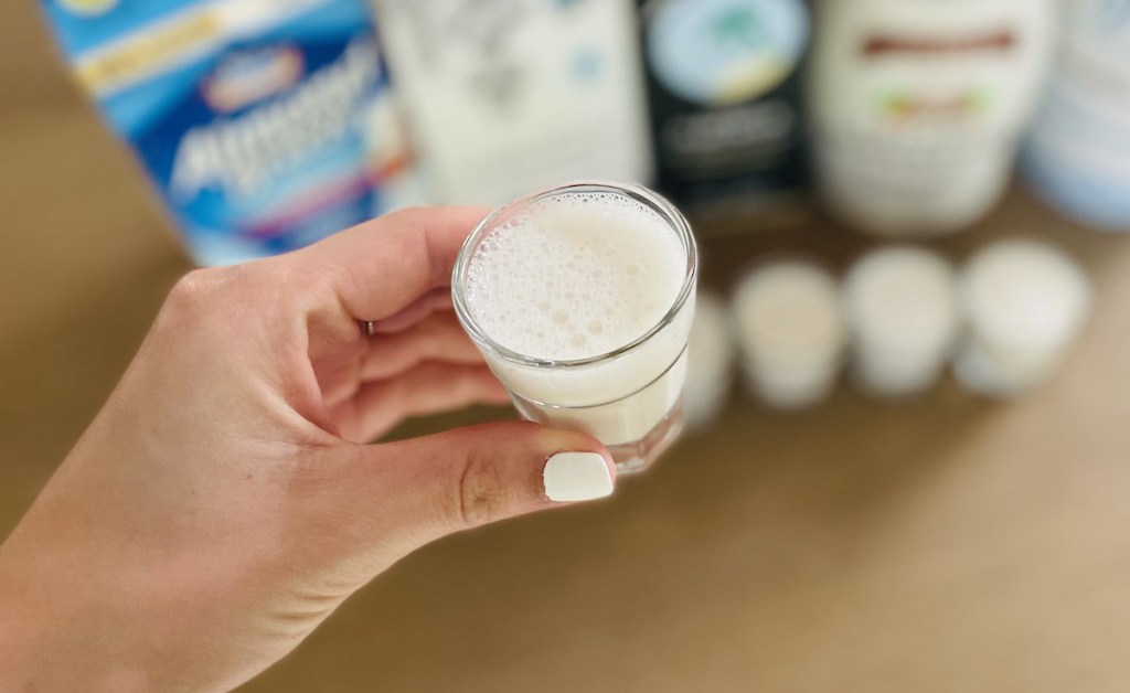 hand holding a shot glass of milk with cartons and bottle of keto dairy free milk in background