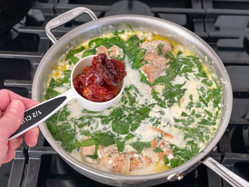 holding a measuring cup of sun-dried tomatoes over a skillet with chicken and cream sauce 
