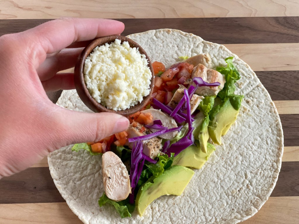 adding cotija to a keto tortilla with lettuce, cabbage, avocado, chicken, and pico de Gallo.