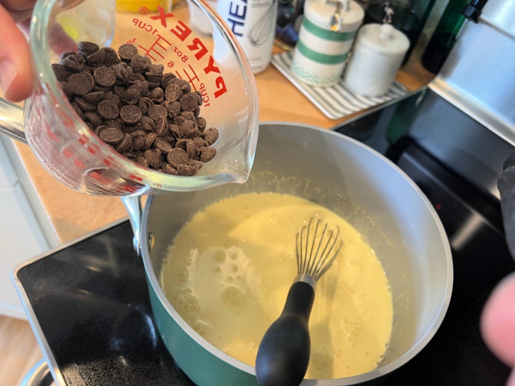 Chocolate chips being poured into a pot with pudding mixture on stovetop