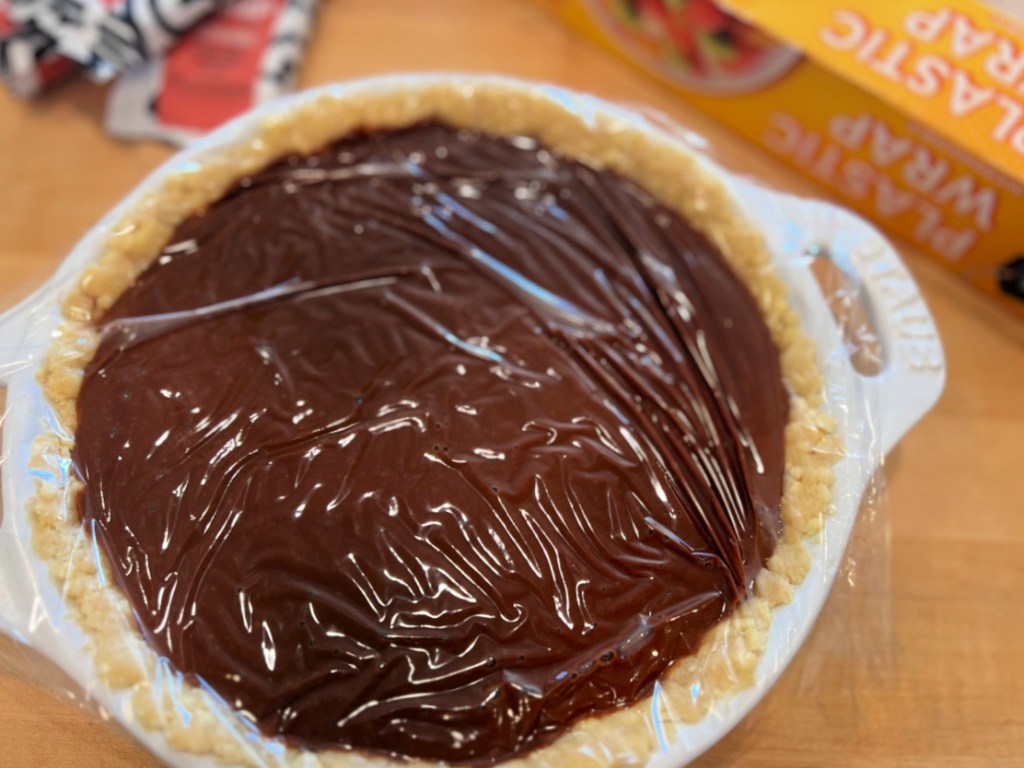 Chocolate pie with pudding layer covered in plastic wrap getting ready to go into refrigerator