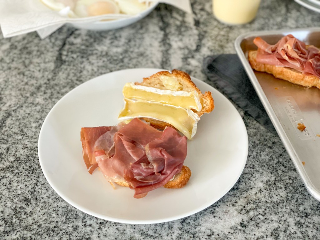 plate with a hero bread croissant cut in half and topped with proscuitto and brie