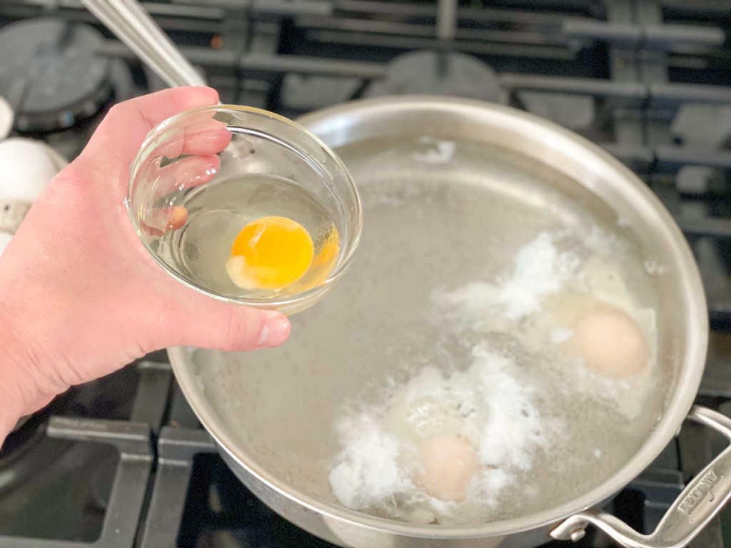 holding a cracked eggs in a small bowl over a skillet with boiling water to poach it