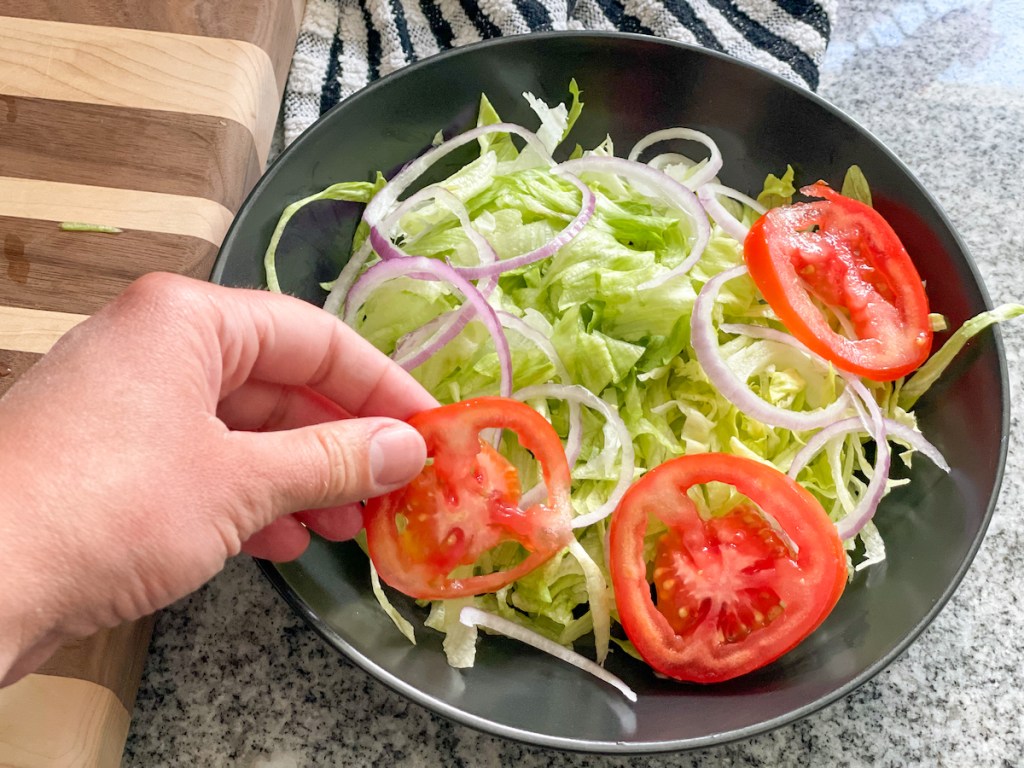 adding tomato slices to a bowl with iceberg lettuce