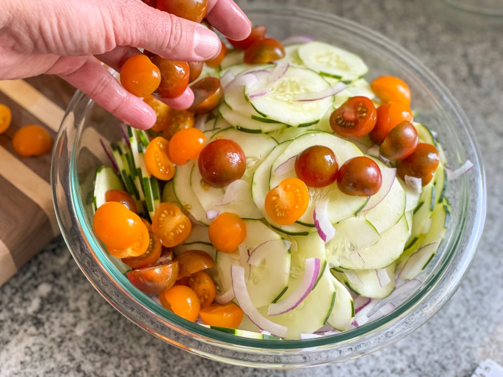 adding sliced cherry tomatoes to a bowl of sliced cucumbers and red onions 