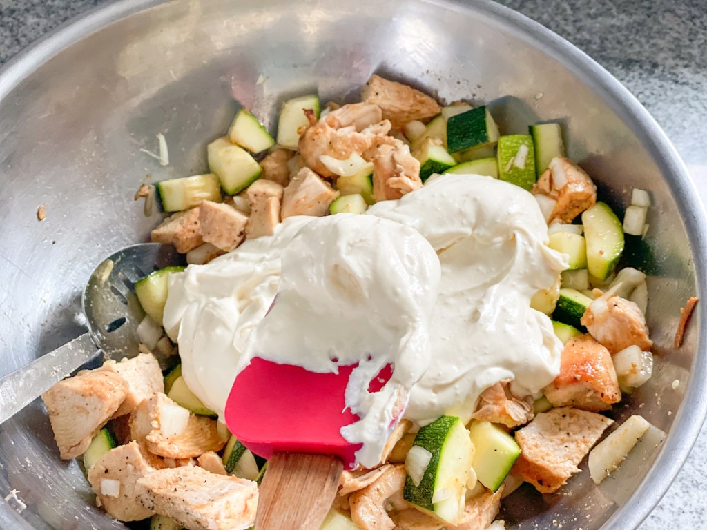 adding a cream sauce to a bowl with chopped chicken, zucchini, and onion